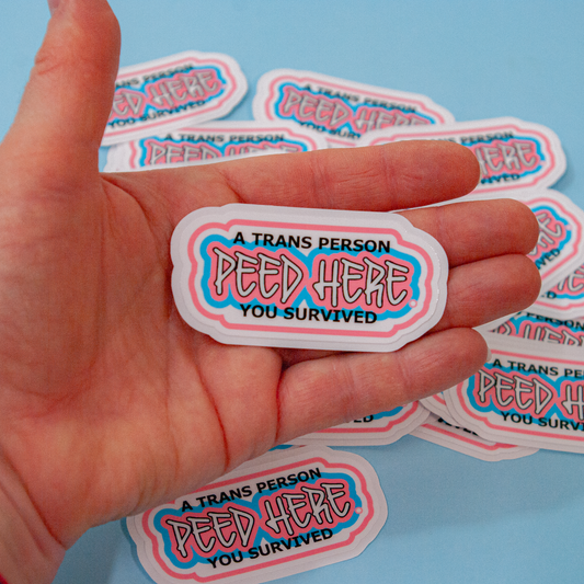 A hand holding a sticker with the text: 'A trans person peed here. You survived.'
