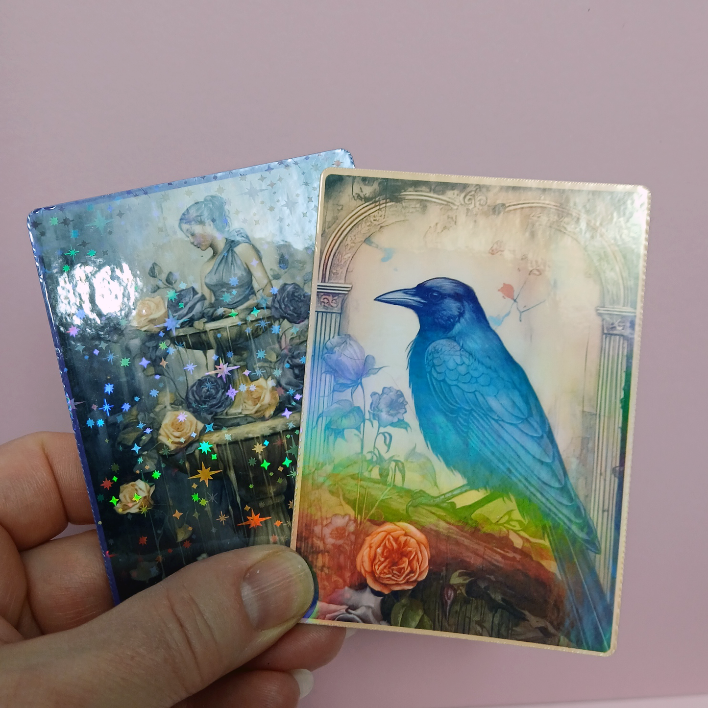 Two TCG cards held by a hand against a pink background, one with a blue bird and the other with a woman and flowers. One card has Holographic stars laminate and card with bird is laminated with Clear holographic laminate